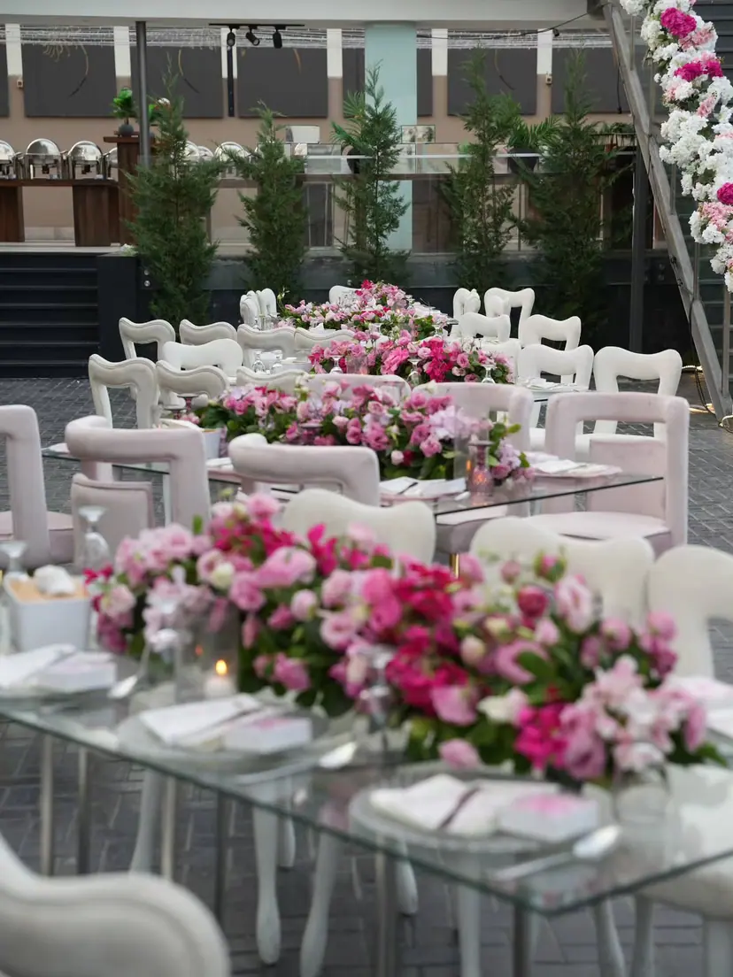 Glass tables lined with pink roses and white chairs at an outdoor wedding