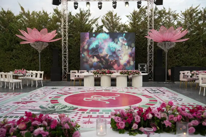 An outdoor wedding stage framed by giant pink lotus sculptures, with a hand-painted monogram dance floor and pink roses