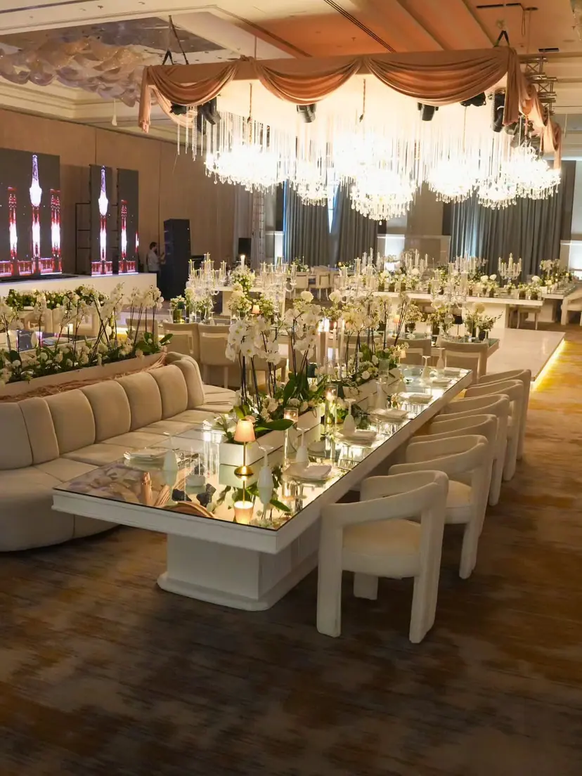 A hotel ballroom set with long mirrored tables, cream sofas and crystal chandeliers