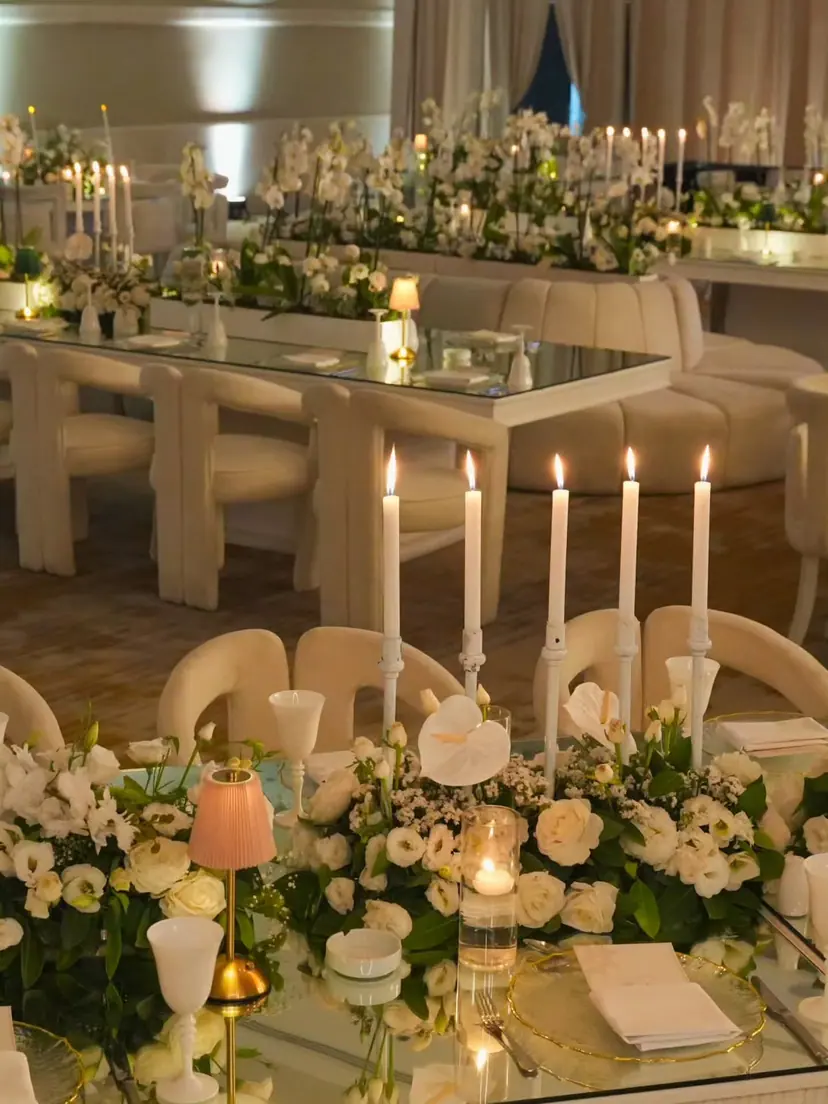 Tall white candles and low white flowers along a mirrored table lined with cream chairs